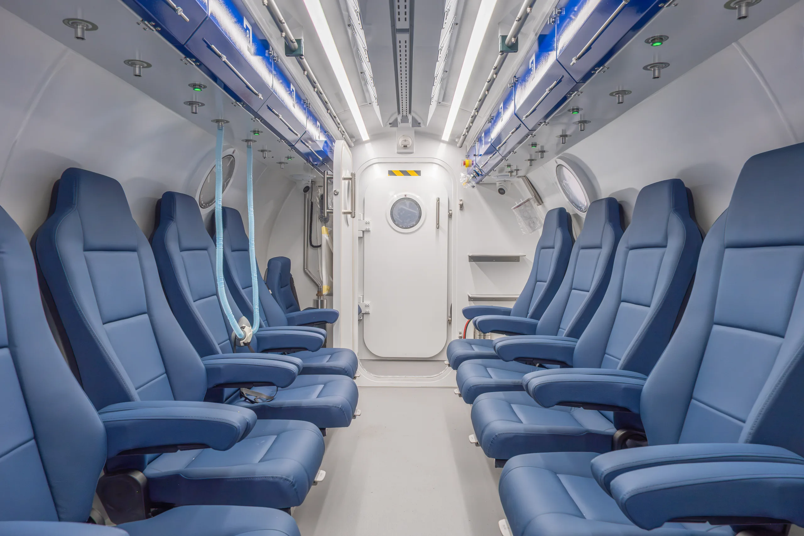 The interior of a Hipertech Multiplace Omega in Athens, seen straight down the aisle: two rows of blue seats either side, round viewports in the white hull, and the inner door with its round window at the far end